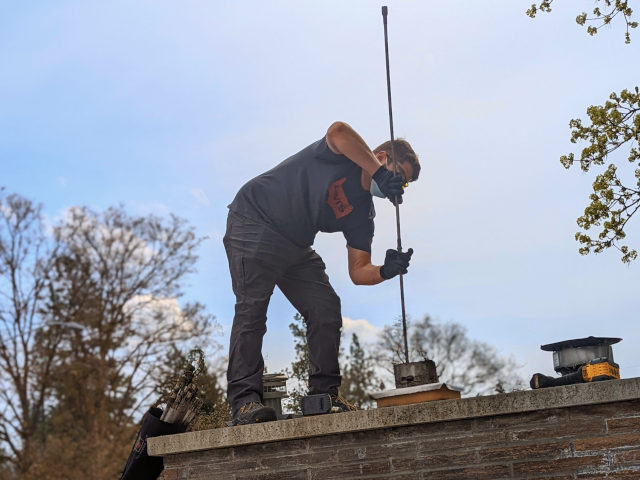 A person standing on top of a masonry chimney is leaning over using rods and a brush to sweep a chimney